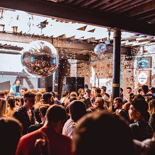 Image taken of dancefloor filled with people inside of nightclub 24 Kitchen Street. There is a DJ, mirror ball and bright lights.